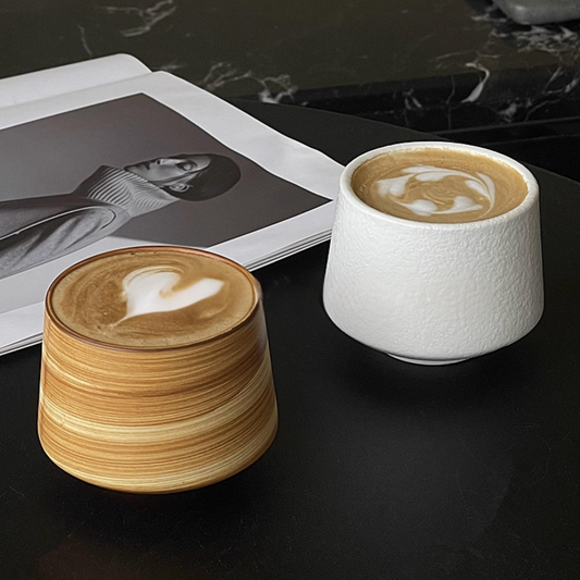 Two ceramic coffee cups with latte art on a dark surface, one wooden and one white.